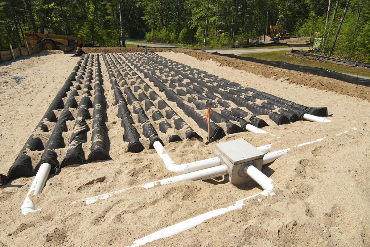 New leach field installation behind a home, showing chambers and distribution manifold with excavation equipment in background